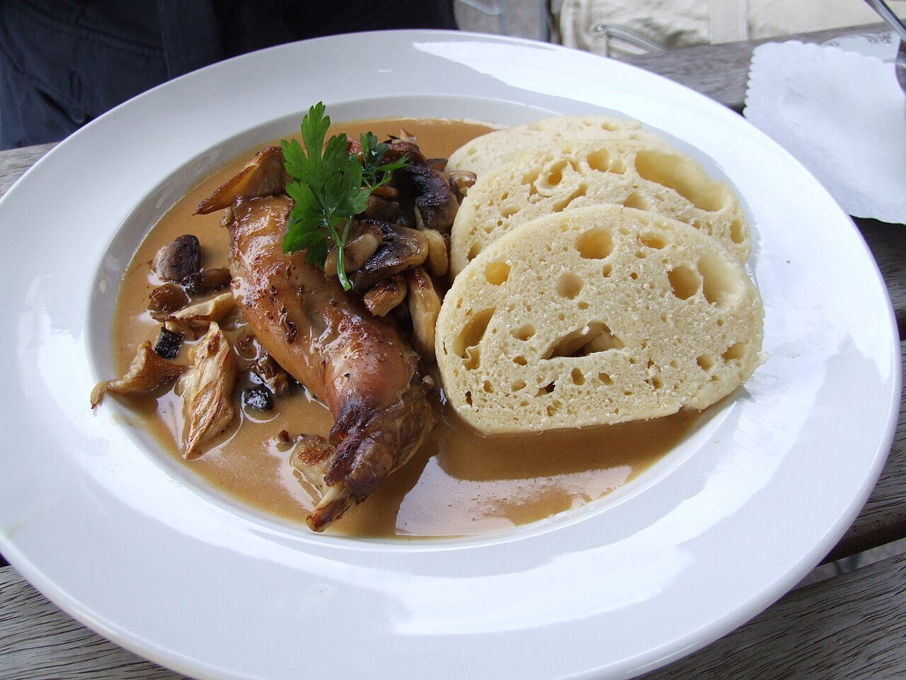 Zdjęcie posiłku na talerzu w postaci mięsa w sosie, obok którego leżą plastry knedliczków, przypominające grube plastry jasnego pieczywa.