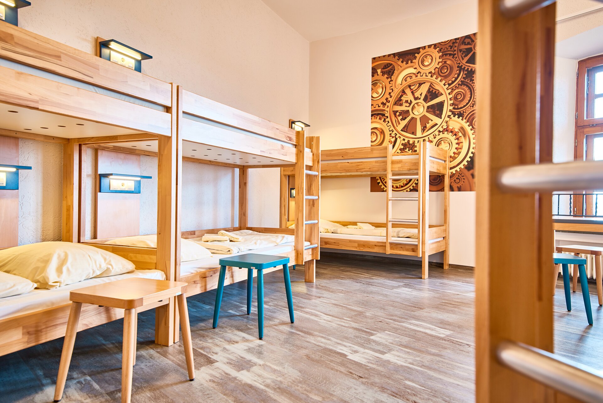 Zdjęcie przedstawia wnętrze pokoju, w którym znajdują się trzy drewniane łóżka piętrowe. Pierwszy plan zajmuje fragment drabinki kolejnego łóżka. Przy łóżkach stoją taborety. Przy każdym łóżku jest zamontowana lampa.