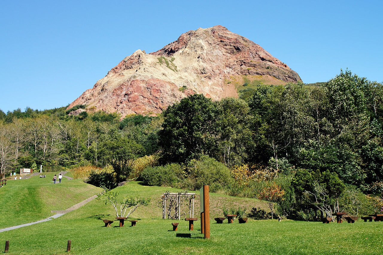 Na zdjęciu znajduje się skaliste wzniesienie. W niektórych miejscach skały mają kolor pomarańczowy. U podnóża góry teren jest zielony - rośnie trawa i drzewa. Jest ścieżka, stoją drewniane ławki. W oddali przechadzają się ludzie. Na fotografii niebo jest bezchmurne.     