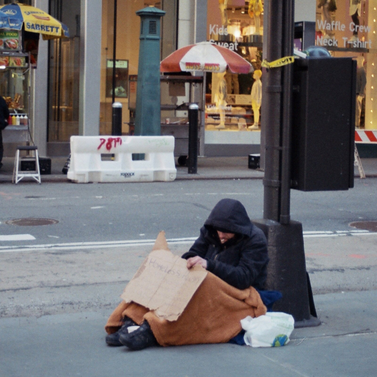 Zdjęcie ukazuje chodnik i ulicę w mieście. Na chodniku osoba w zimowej kurtce, z kapturem naciągniętym na głowę, siedzi przykryta kocem. Człowiek przedstawiony na fotografii opiera się plecami o metalowy słup. Transkrypcja nagrań znajduje się poniżej.