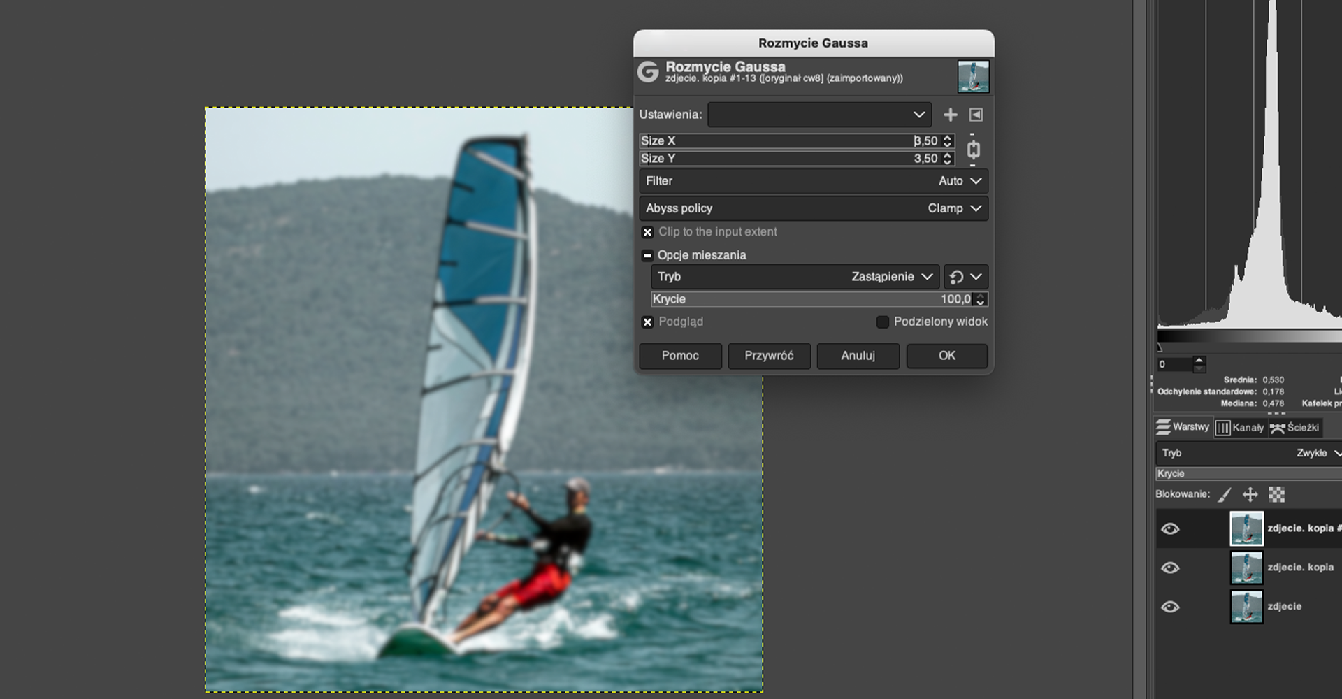 Ilustracja przedstawia okno programu. W obszarze roboczym jest zdjęcie mężczyzny na windsurfingu. Zdjęcie jest rozmyte, nieostre. W obszarze roboczym jest okno dialogowe o nazwie: Rozmycie Gaussa. Zaznaczono Size X 3,50, Size Y 3,50, Filter Auto, Abyss policy - Clamp. Zaznaczono Clip to the input extent. Tryb - Zastąpienie, Krycie 100,0. Zaznaczono: Podgląd. Po prawej stronie jest panel dotyczący warstw. Tam zdjecie.kopia #1, zdjęcie.kopia, zdjecie. Zaznaczono zdjecie.kopia #1.  