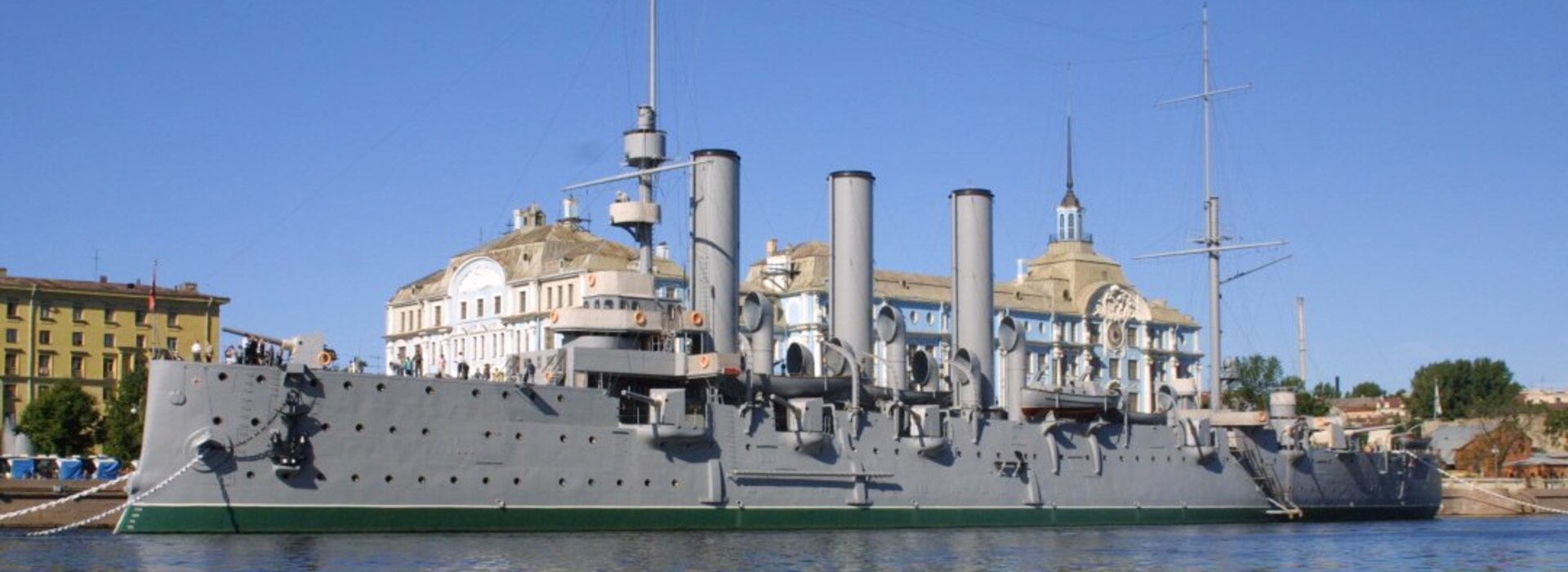 Fotografia barwna przedstawiająca Krążownik "Aurora". Ukazuje duży uzbrojony okręt wojenny w porcie. W tle widoczne są miejskie budynki.