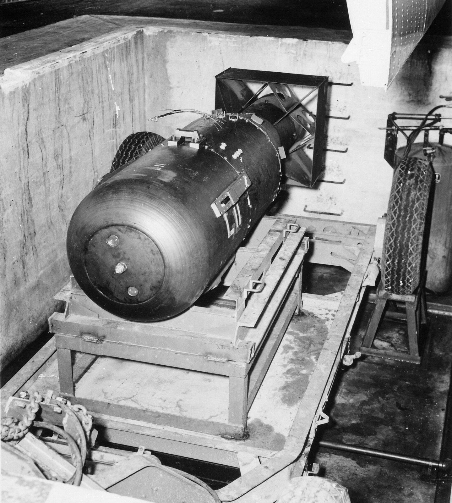 Czarno‑białe zdjęcie 1: bomba atomowa Little Boy zrzucona na Hiroszimę. Bomba w kształcie cylindra. Długość około 1 metr, średnica około 50 centymetrów. Bomba umieszczona jest na metalowej ramie w kształcie prostopadłościanu.