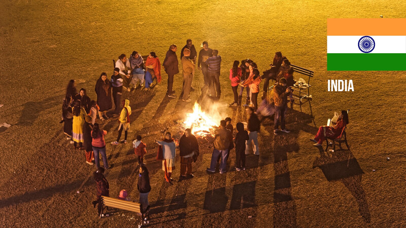 Zdjęcie przedstawia dużą grupę osób, które stoją wokół zapalonego ogniska, na płaskim terenie porośniętym trawą. Część z osób prowadzi rozmowy w mniejszych grupkach. Po prawej stronie ogniska i w dolnej części fotografii widoczne są ustawione ławki, na których siedzą uczestnicy ogniska. W prawy boku widoczna jest flaga Indii. 