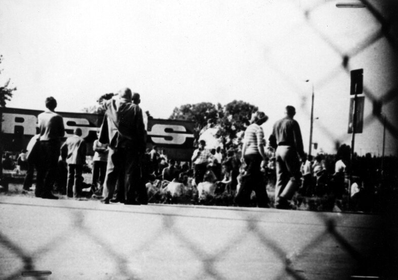 Czarno-biała fotografia przedstawiająca strajk w ZM “Ursus” w 1976 roku. Liczna grupa robotników jest zgromadzona na placu. Zdjęcie wykonano zza płotu z siatki.