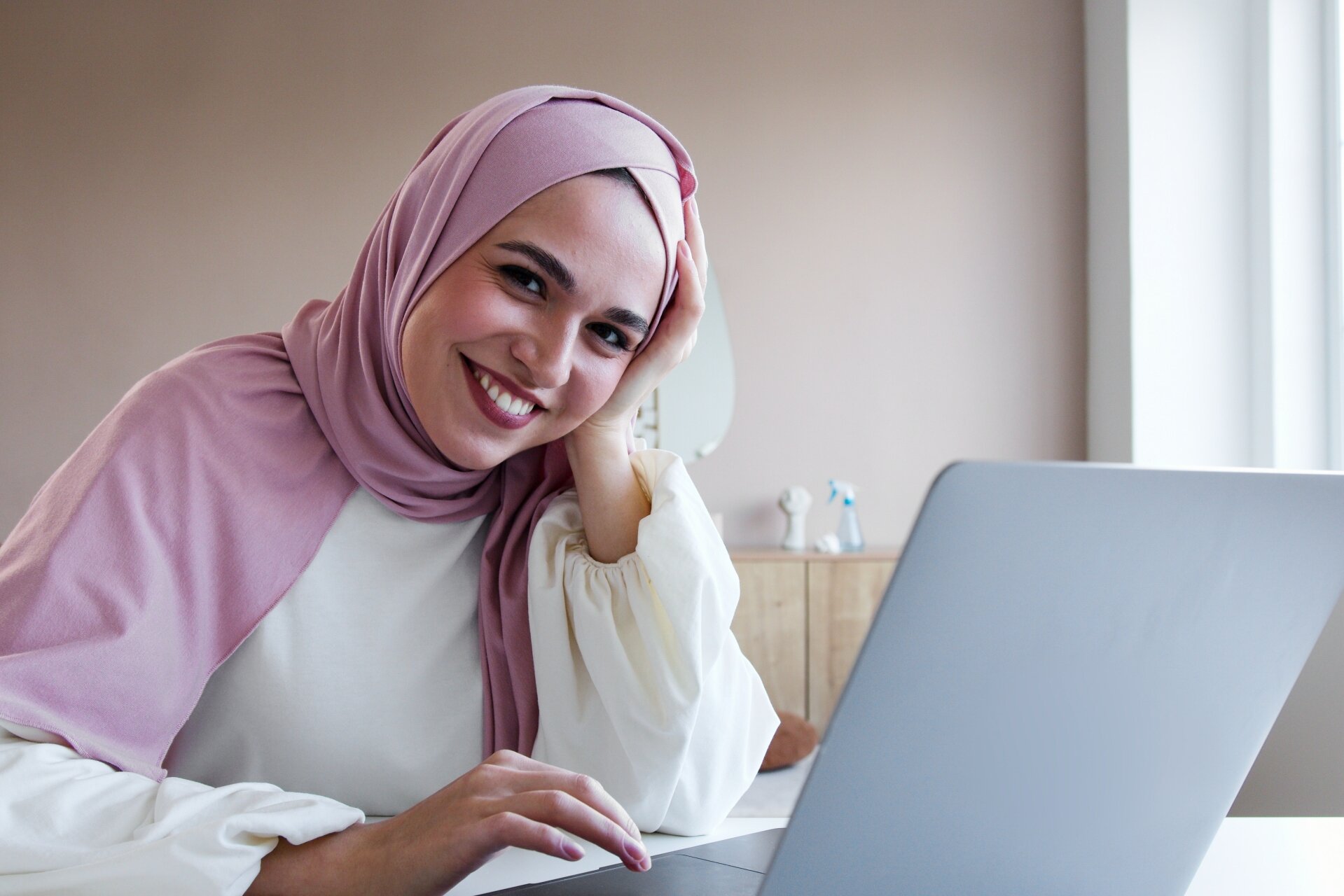 Zdjęcie przedstawia młodą kobietę siedzącą przy stole, przy otwartym laptopie. Kobieta ma ciemną oprawę oczu, uśmiecha się szeroko. Ubrana jest w białą bluzkę.Na głowę i ramiona ma nałożoną dużą chustę. 