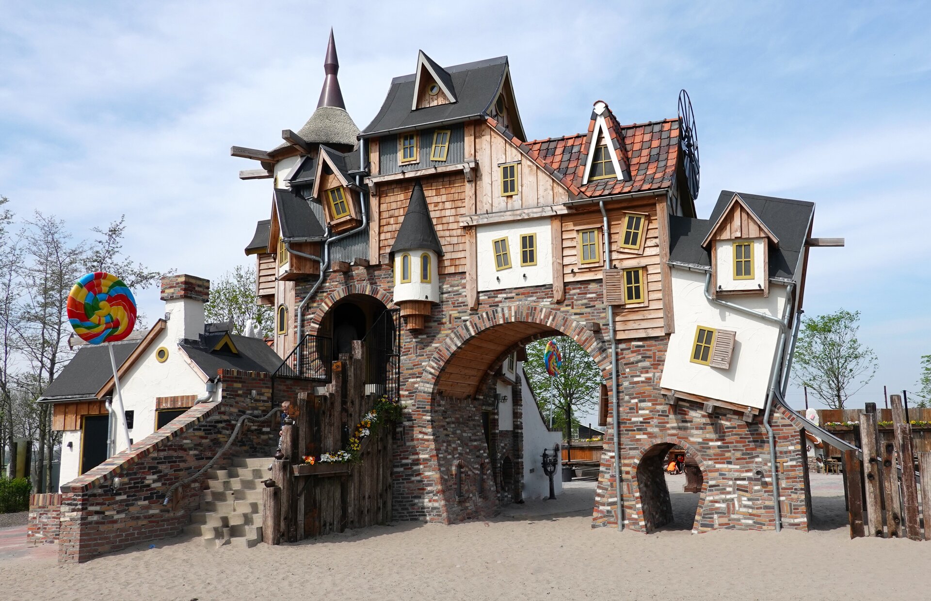 Zdjęcie przedstawia fragment muru z bramą przejściową, wokół którego jest pusta przestrzeń. Górną część muru tworzą wklejone w niego budynki ustawione pod różnymi kątami odbiegającymi od pionu. Każdy z budynków jest w innym stylu, np. murowany, biały piętrowy fragment jest tuż obok elementu z drewnopodobną elewacją i użytkowym poddaszem.