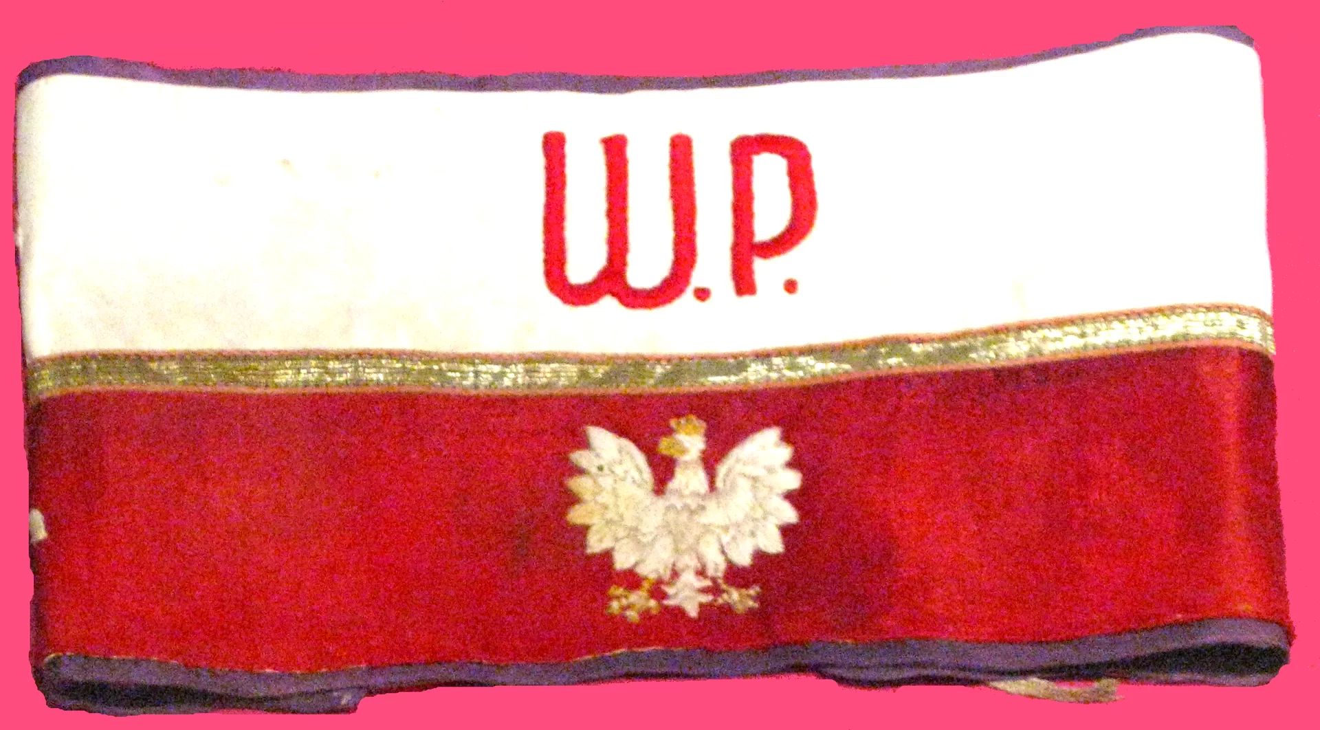 Kliknij, aby powiększyć Zdjęcie przedstawia opaskę. Ma dwie narodowe barwy - białą i czerwoną. Na białej wyszyte są litery: W.P. Na czerwonej - wyszyty jest orzeł. Patrzy w lewo, nosi złotą koronę na głowie.