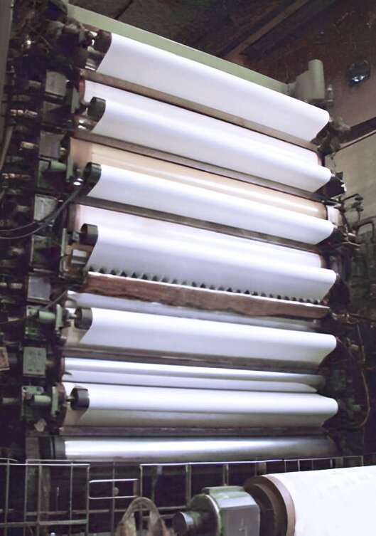 Grafika przedstawia kalander. Jest to wysoka maszyna składająca się z wielu walców umieszczonych jeden nad drugim.