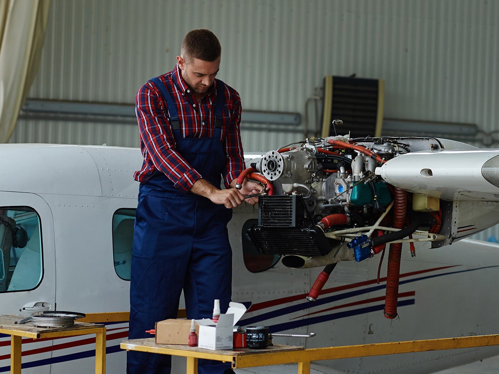 Zdjęcie przedstawia młodego mężczyznę, który stoi na podnośniku i naprawia mechanizm skrzydła samolotu. Mężczyzna jest ubrany w kraciastą koszulę i granatowe robocze spodnie na szelkach. W prawej dłoni trzyma jakie narzędzie, którym dotyka elementu mechanizmu skrzydła, a lewą dłonią przytrzymuje czerwony, gruby przewód. Przed nim widoczne są dwa małe blaty, na których leżą narzędzia, akcesoria i kable. W tle za nim widoczny jest kadłub samolotu znajdującego się w hangarze.