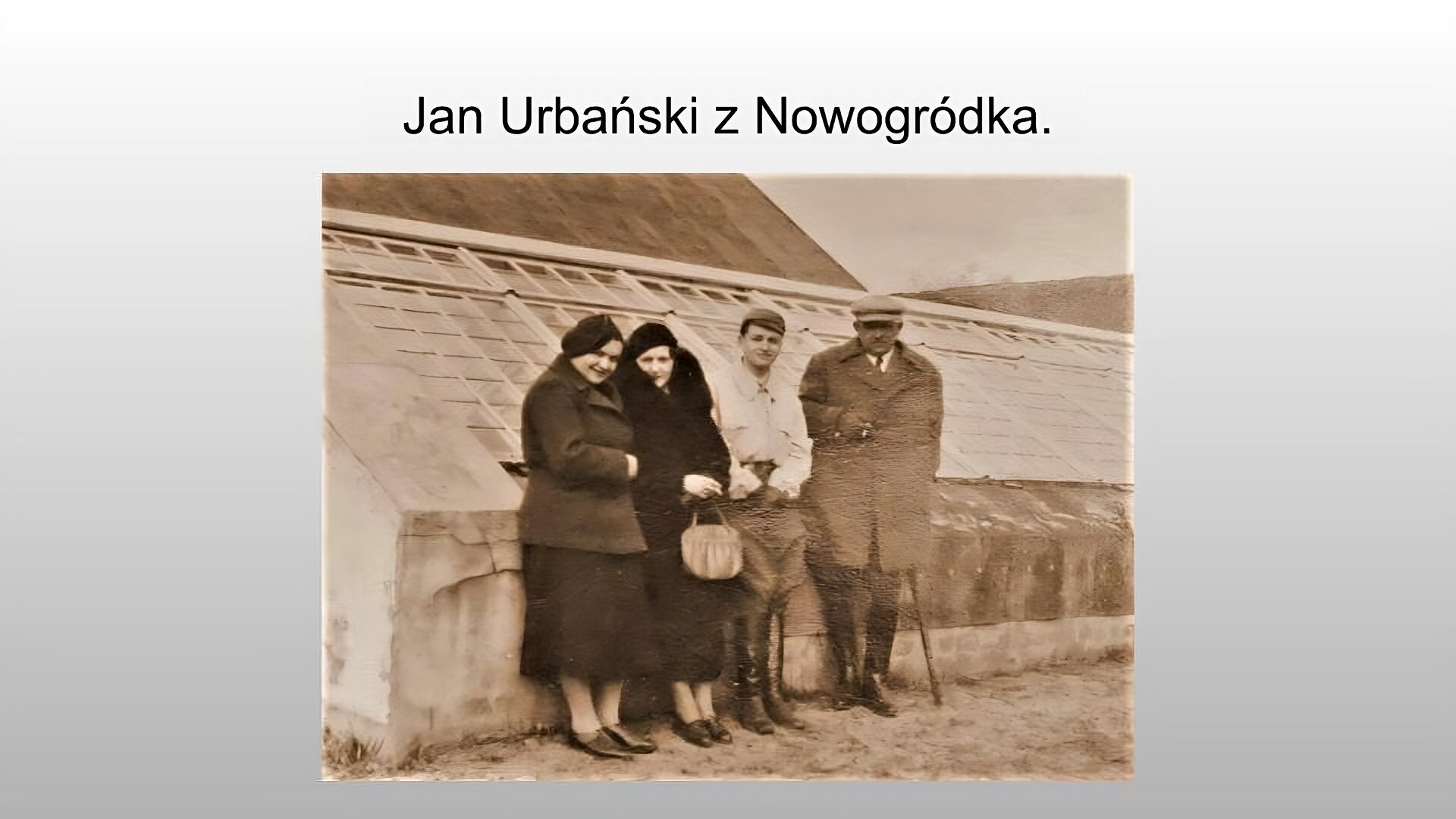 Jasnoszary slajd. Na górze slajdu napis: „Jan Urbański z Nowogródka”. Poniżej zdjęcie przedstawiające cztery osoby stojące na tle niskiej szklarni. Pierwsza osoba od lewej, kobieta, ubrana jest w długą spódnicę za kolana, skórzane niskie buty, grubą kurtkę i beret. Kobieta uśmiecha się. Obok niej stoi druga kobieta, ubrana w niskie, skórzane buty, płaszcz i beret. W ręku trzyma małą, jasną torebkę. Obok niej stoi młody mężczyzna w wysokich, sięgających do kostek, butach skórzanych, luźnych spodniach, białej koszuli i czapce. Obok niego stoi czwarta postać, starszy mężczyzna. Ma on wysokie, skórzane buty, luźne spodnie wpuszczone w buty, długi płaszcz. Na głowie ma czapkę, prawą dłoń trzyma na guziku płaszcza, a lewą podpiera na lasce. Zdjęcie jest sepią.