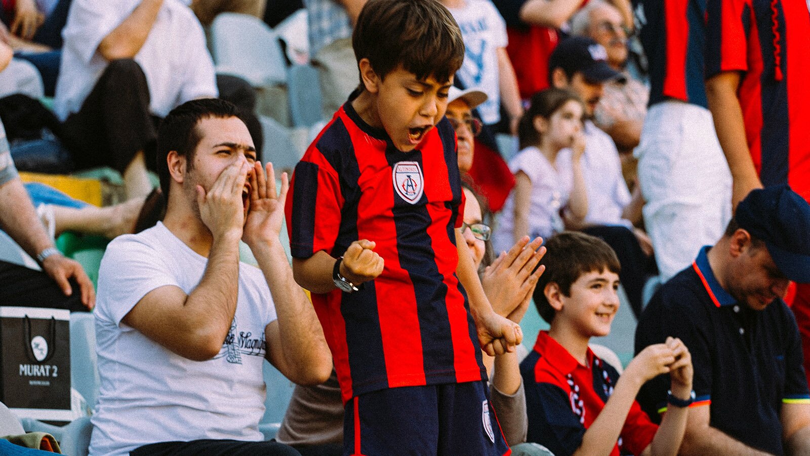Zdjęcie przedstawia kibiców na trybunach. Na pierwszym planie widoczny jest chłopiec, ubrany w koszulkę z krótkim rękawem w pionowe czarne i czerwone paski. Chłopiec stoi, prawą rękę ma zgiętą, a dłoń zaciśniętą w pięść, zaś lewą rękę ma opuszczoną, a dłoń również zaciśniętą. Po obu stronach chłopca widoczne są osoby, które krzyczą i klaszczą.