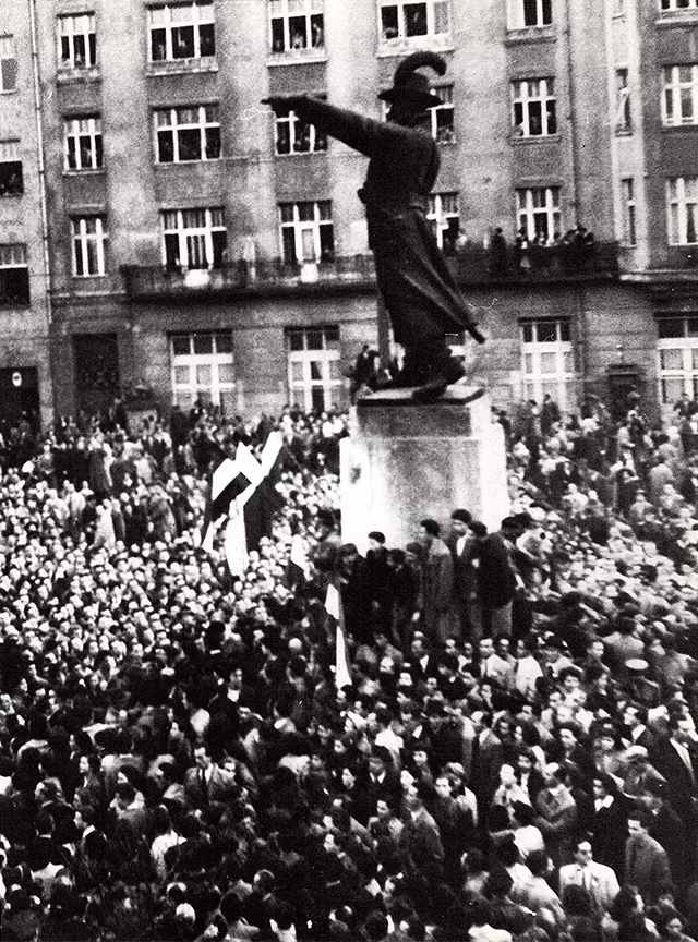 Zdjęcie przedstawia tłum ludzi manifestujący na ulicy. Na środku stoi pomnik.