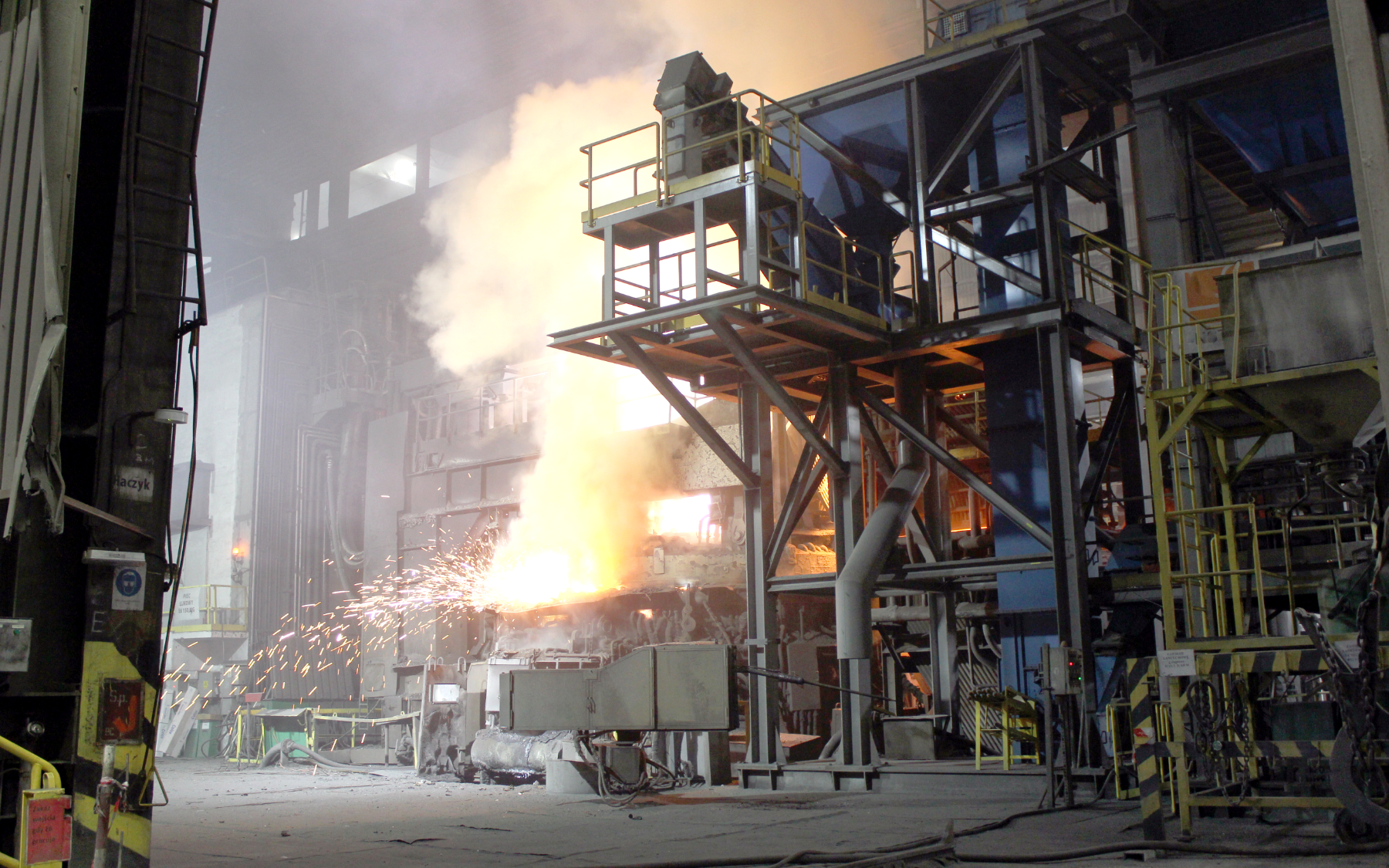 Ilustracja przedstawia halę zakładu odlewniczego, w którym realizowany jest proces wytopu stali w elektrycznym piecu łukowym. Widoczne są złożone i wysokie konstrukcje stalowe, z pomiędzy których strzelają słupy iskier. Źródłem iskier i dymu jest piec łukowy.