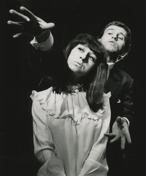 Czarno‑białe zdjęcie przedstawia aktorkę i aktora w trakcie wystąpienia. Z przodu siedzi kobieta w jasnej, bufiastej bluzce. Ma długie włosy z grzywką  i lekko pochyloną głowę w prawą stronę. Za nią stoi mężczyzna w garniturze. Ma krótkie włosy i lekko zarysowany zarost. Wskazuje swoją prawą ręką kierunek przed siebie. Kobieta patrzy w tę stronę.