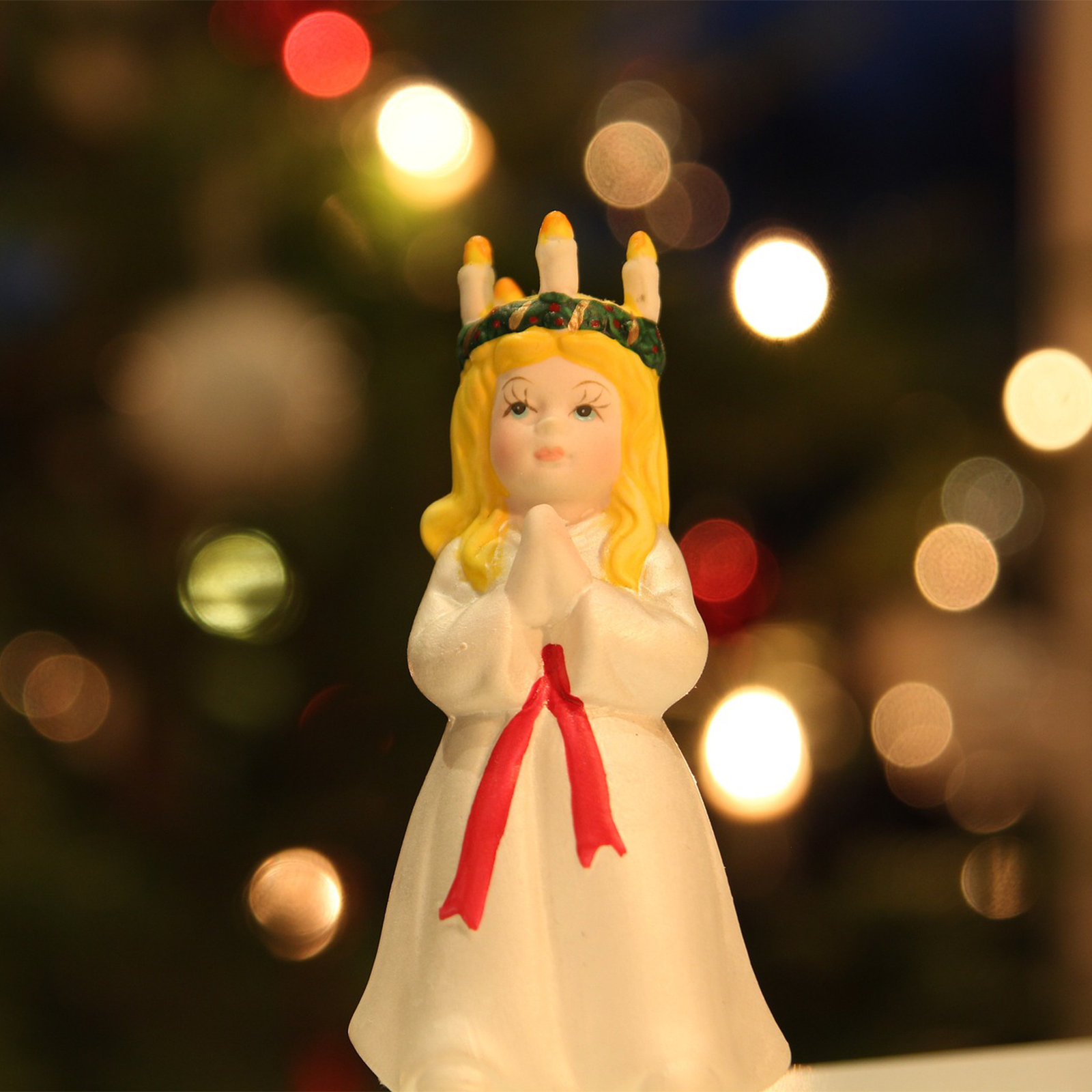 Zdjęcie przedstawia figurkę dziewczyny z długimi włosami w kolorze żółtym, ubraną w długą białą sukienkę z czerwoną szarfą w pasie. Figurka ma ręce złożone jak do modlitwy. Na jej głowie  widoczny jest wieniec ze świeczkami. 