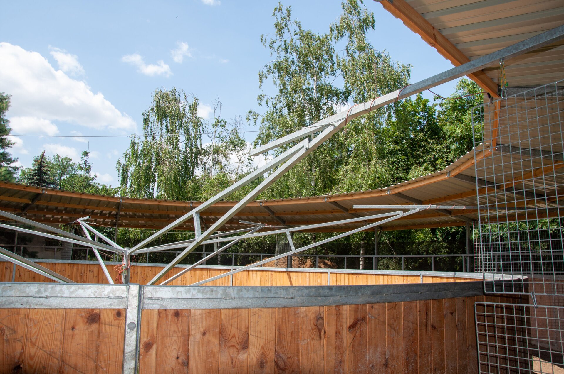 Widok na karuzelę boksową. Ma ona okrągły kształt. Boki i dach wykonane są z drewna, natomiast konstrukcja obrotowa z siatkowymi przegrodami wykonana jest ze stali. Dach osłania tylko tor, po którym porusza się koń, środek karuzeli jest niezadaszony.