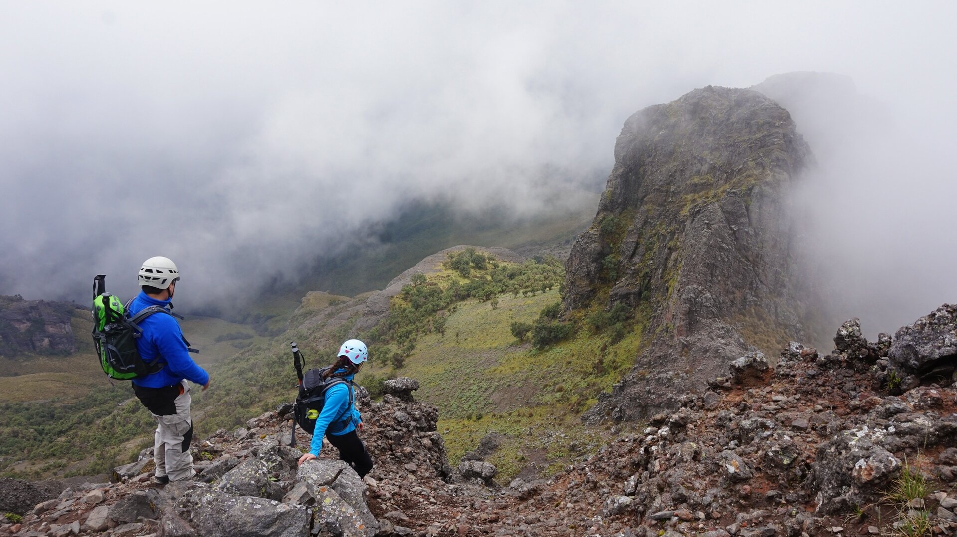 Na zdjęciu znajduje się dwoje turystów w trakcie pieszej wędrówki w górach, schodzących ze skały w dół. Na głowach mają kaski ochronne, na plecach niosą plecaki. Góry otoczone są mgłą.