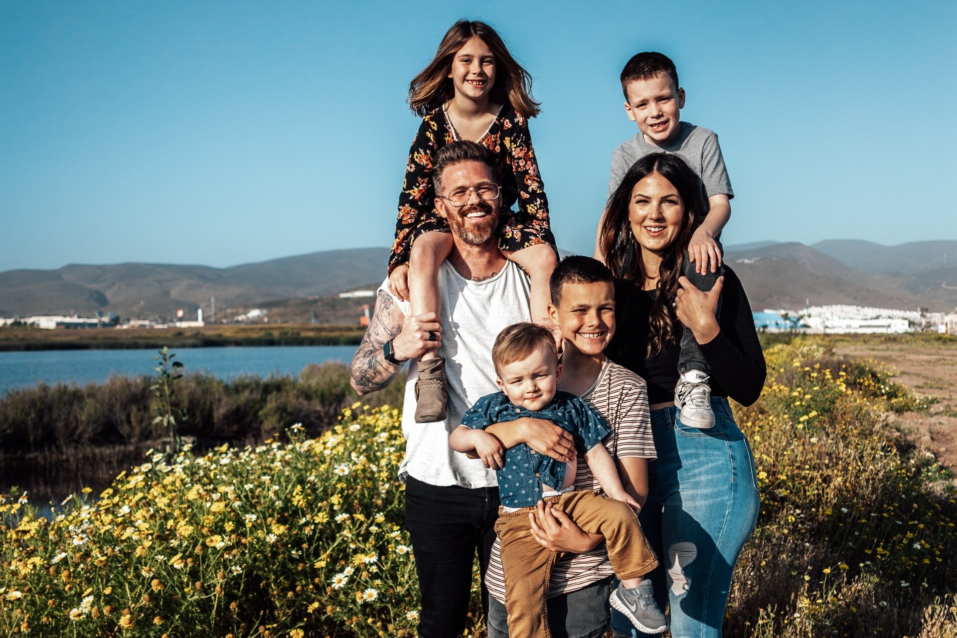 Ilustracja przedstawia mężczyznę, kobietę i czworo dzieci, jedną dziewczynkę i trzech chłopców, tworzących uśmiechniętą rodzinę. Mężczyzna i kobieta trzymają na plecach dzieci, a starszy chłopiec stoi pośrodku i opiekuje się najmłodszym. W tle widoczne są wody jeziora i łagodne wzniesienia tworzące spokojną scenerię wypoczynku na łonie natury.