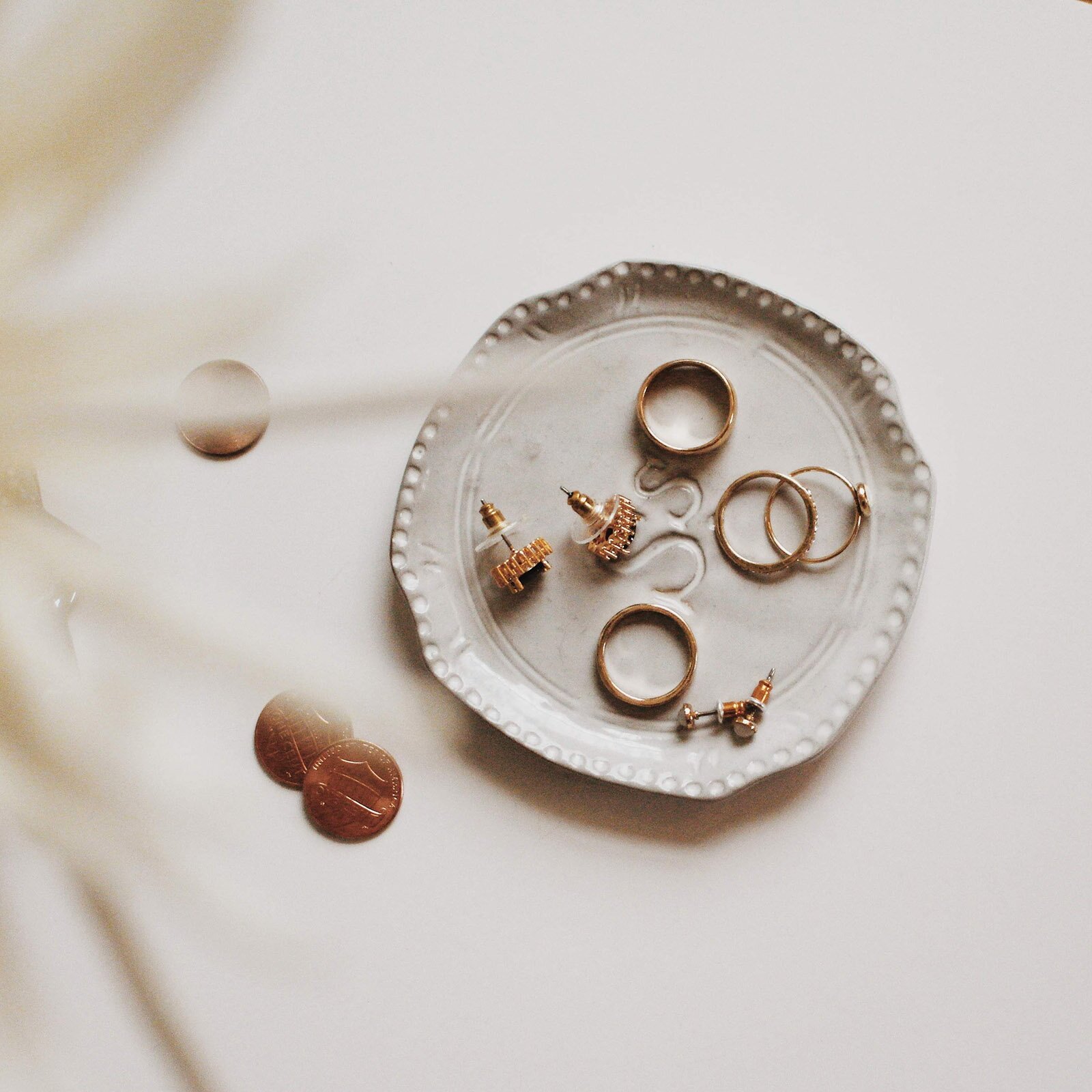 Zdjęcie przedstawia dekoracyjny, jasnoszary, porcelanowy talerzyk, na którym leżą złote pierścionki, obrączki oraz dwie pary kolczyków. Po lewej stronie talerzyka widoczne są trzy monety.