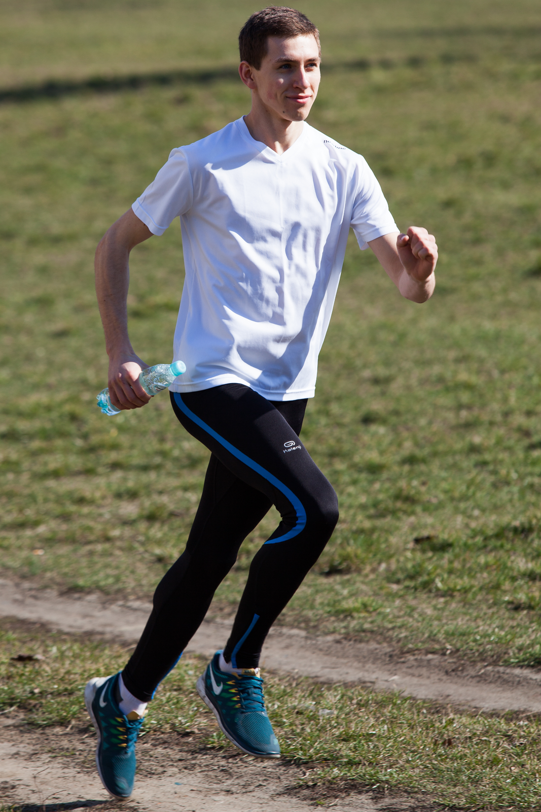 Fotografia przedstawia uśmiechniętego, biegnącego w prawo człowieka. Ubrany w białą koszulkę, czarne spodnie z niebieskimi wzorami, niebieskie bury sportowe. W ręce trzyma butelkę wody.