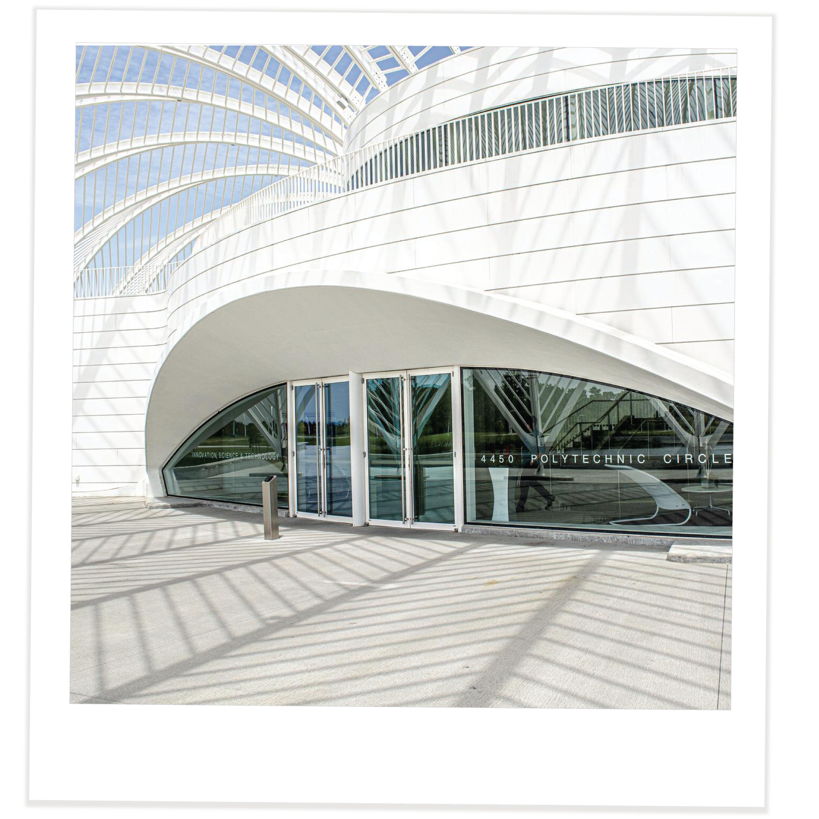 Zdjęcie przedstawia budynek o nowoczesnej architekturze. Jest biały i ma zaokrąglone kształty. Z przodu przeszklone wejście z napisem na szybie POLYTECHNIC CIRCLE.