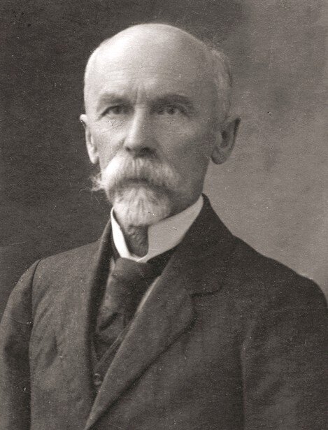 Zdjęcie przedstawia starszego, łysego mężczyznę w garniturze. Ma gęste, siwe wąsy oraz małą brodę. Ma zmarszczki na twarzy.