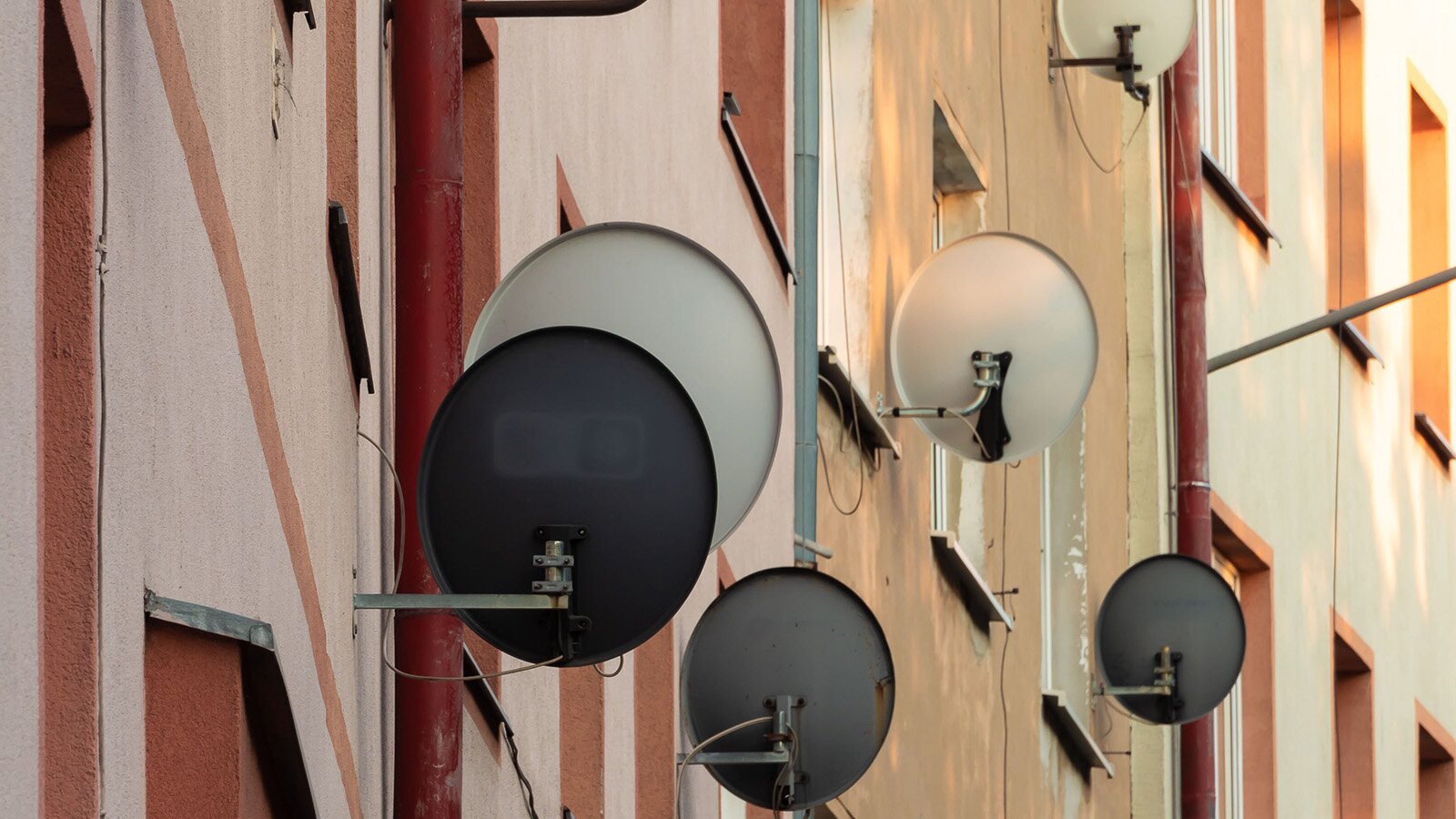 Zdjęcie przedstawia z ukosa ścianę budynku wielomieszkaniowego. Na ścianie widoczne są zamontowane przy oknach anteny satelitarne.
