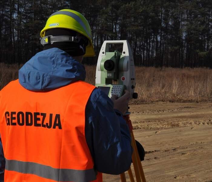 Zdjęcie przedstawia mężczyznę w kamizelce odblaskowej i kasku stojącego przy teodolicie. Jest to prostokątne urządzenie pomiarowe stojące na trójnogu.