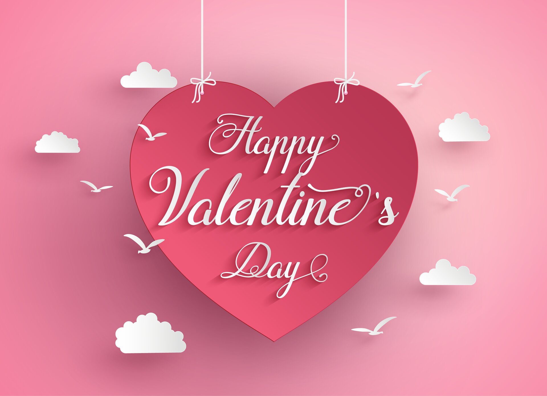 Ilustracja przedstawia typową kartkę walentynkową. Na różowym tle znajduje się czerwone serce wiszące na dwóch białych nitkach, ozdobione białymi chmurkami i ptaszkami. Na sercu widoczny jest biały napis: Happy Valentine s Day.