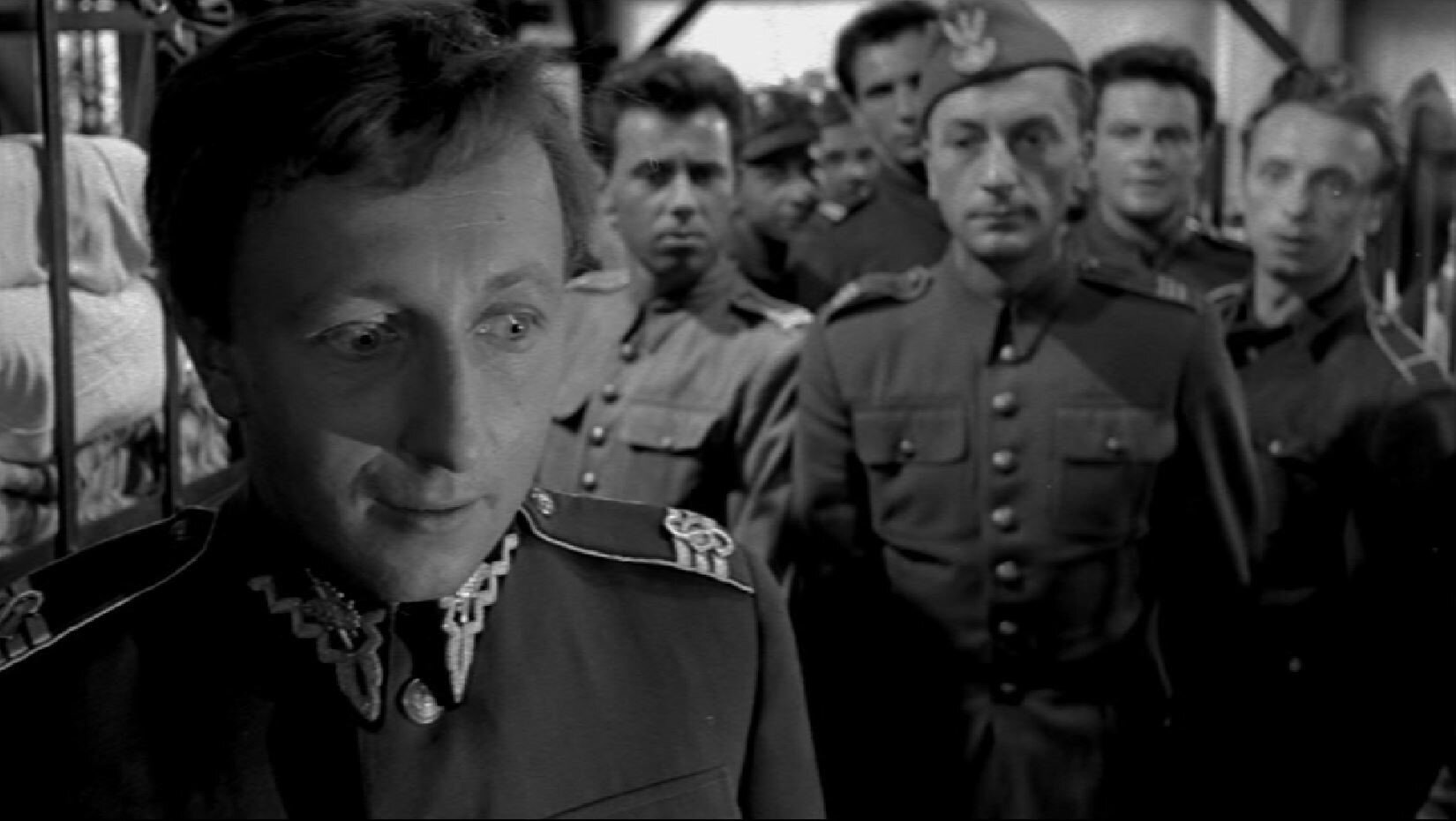 Zdjęcie przedstawia salę z piętrowymi pryczami. Na pierwszym planie znajduje się mężczyzna w mundurze polskiego wojskowego. Za nim, w niewielkim oddaleniu, inni mężczyźni w mundurach polskich wojskowych.