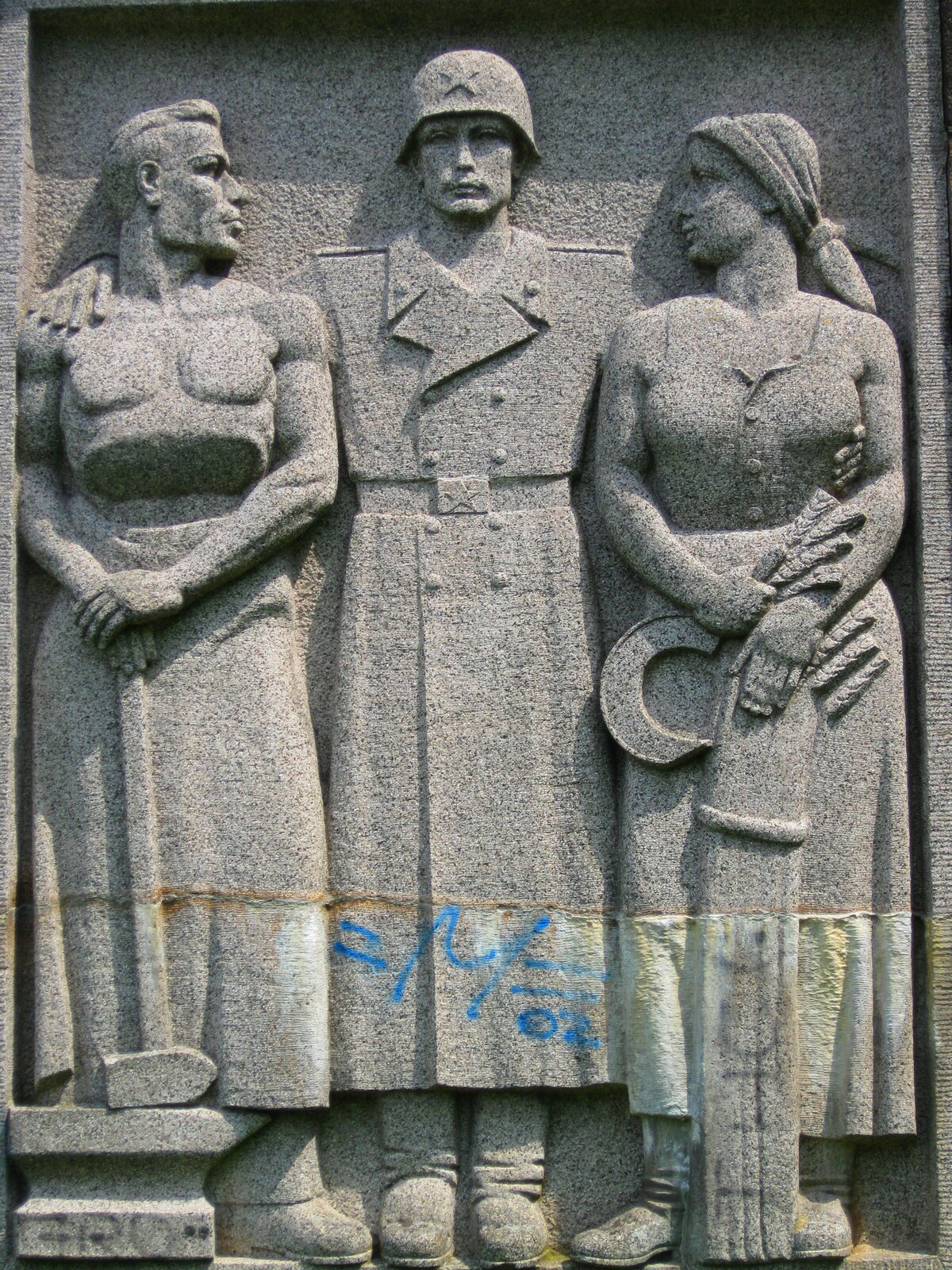 Zdjęcie przedstawia płaskorzeźbę. Widoczne są na niej trzy postacie. Pośrodku stoi radziecki żołnierz ubrany w wojskowy płaszcz, na głowie ma hełm. Obejmuje swoim lewym ramieniem młodą kobietę, trzymającą w dłoniach sierp, prawym ramieniem mężczyznę z młotem w dłoni.