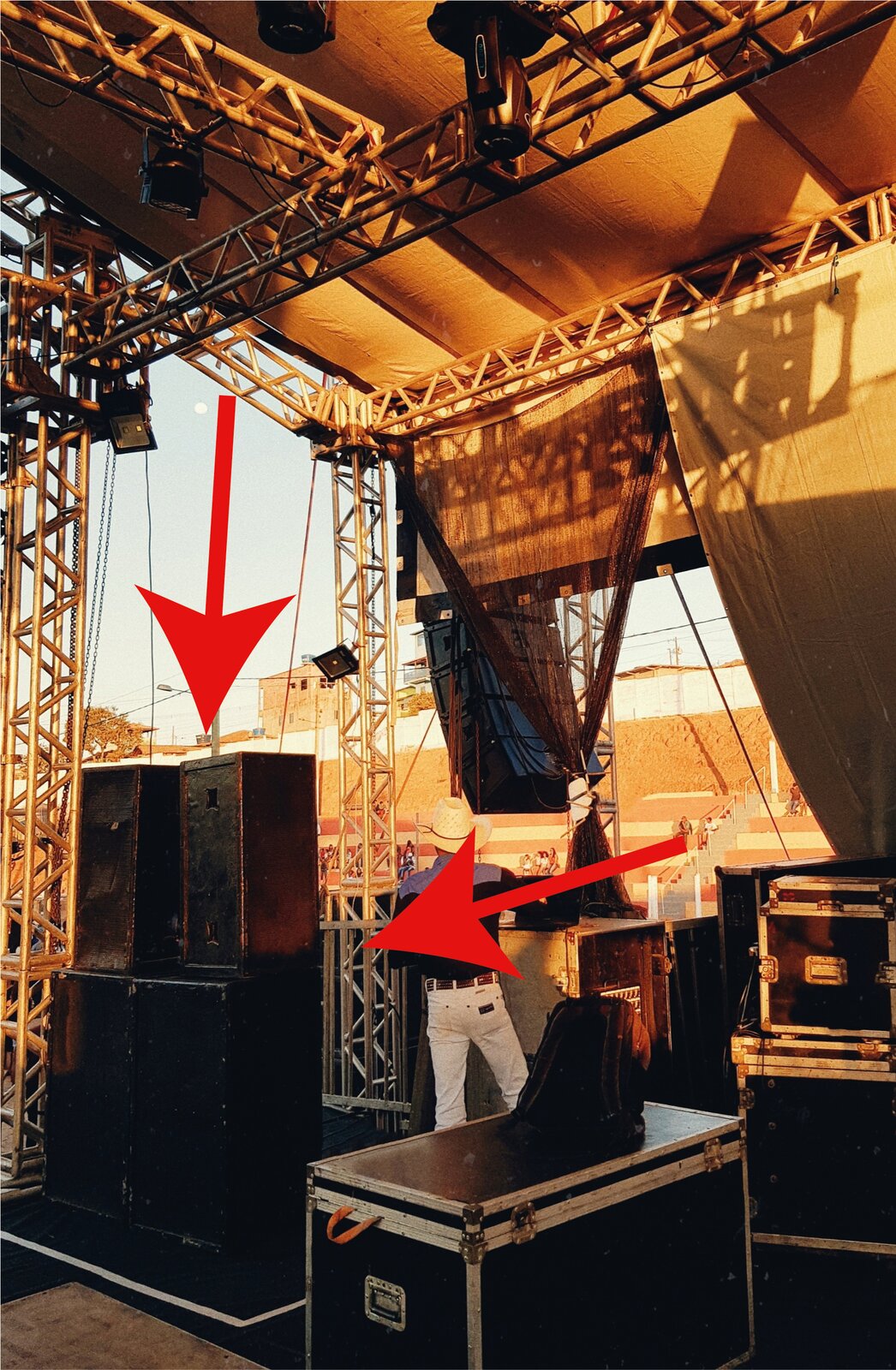 Zdjęcie przedstawia instalację nagłośnienia liniowego podwieszanego, line‑array oraz scenę koncertową. Na pierwszym planie widoczny jest czarny prostopadłościan, w ujęciu trzy czwarte, z metalowymi okuciami – skrzynia na głośniki. Na lewo widoczny jest klaster nagłośnieniowy, czyli zestaw głośników ustawionych w pionie. W dolnej części duży, czarny prostopadłościan, na którym ustawione są dwa mniejsze czarne prostopadłościany. Na ten element wskazują dwie czerwone strzałki, jedna biegnąca z prawej strony, pośrodku, druga od góry. U góry widoczne jest rusztowanie w formie krzyżujących się pionowych i poziomych rurek, pomiędzy którymi zygzakowato biegną cieńsze rurki. Po prawej stronie u widoczne są podwieszone tkaniny w postaci ciemnych i jasnych elementów. Pod czerwoną strzałką pośrodku człowiek stojący tyłem.