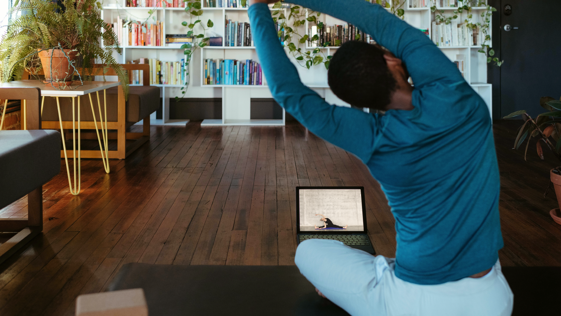 Zdjęcie przedstawia dziewczynę wykonującą ćwiczenia w pokoju. Siedzi tyłem do kadru na macie do ćwiczeń. Trzymając ręce nad głową wykonuje skłon w lewą stronę. Podłoga w pokoju wykonana jest z desek. Przed dziewczyną na podłodze leży otwarty laptop. Na ekranie trener pokazuje wykonanie dokładnie tego samego ćwiczenia. Na dalszym planie w pokoju stoją regały z książkami. Po lewej stronie znajduje się stolik, na którym stoi paproć w doniczce.