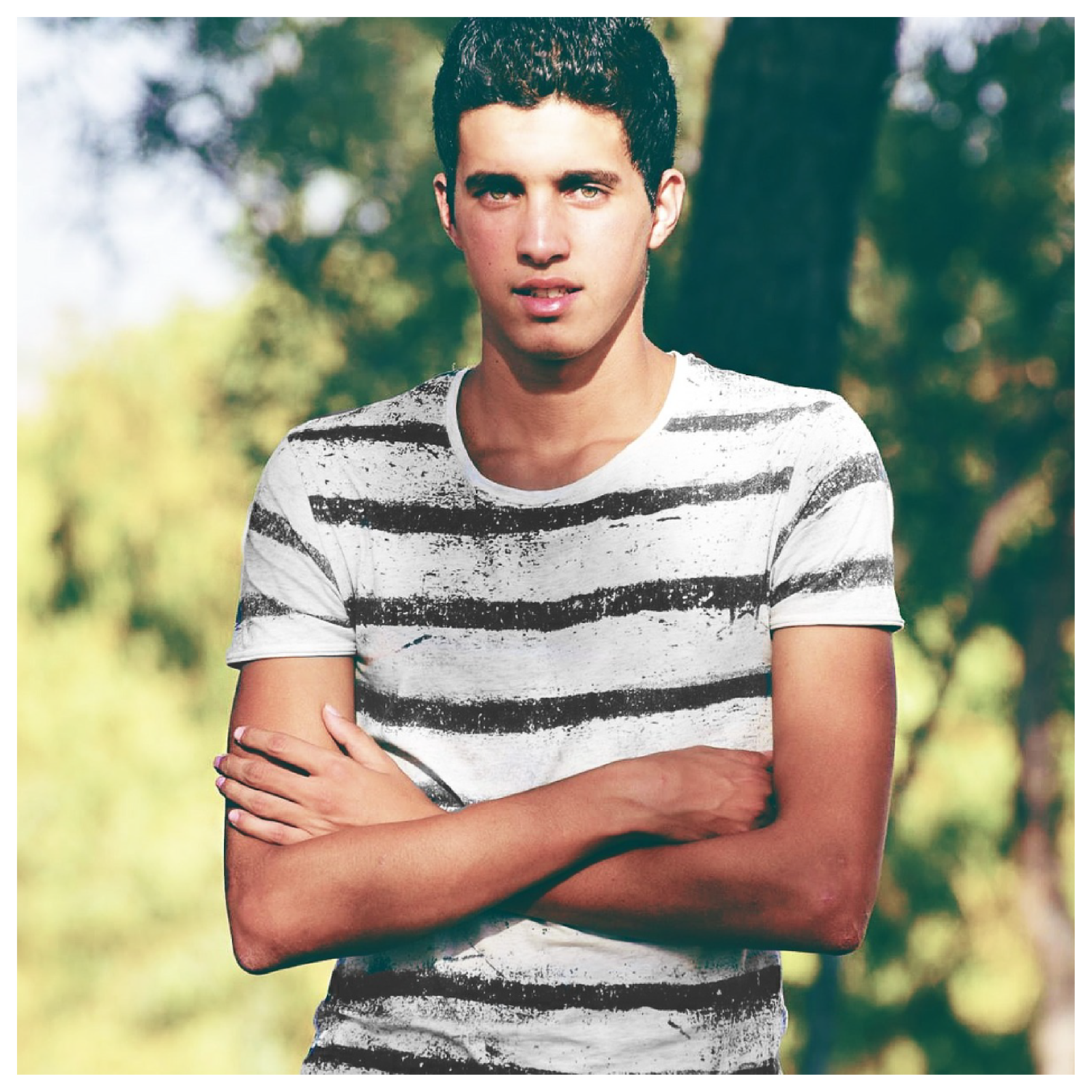 Grafika przedstawia młodego mężczyznę o krótkich czarnych włosach, który stoi z założonymi rękami.