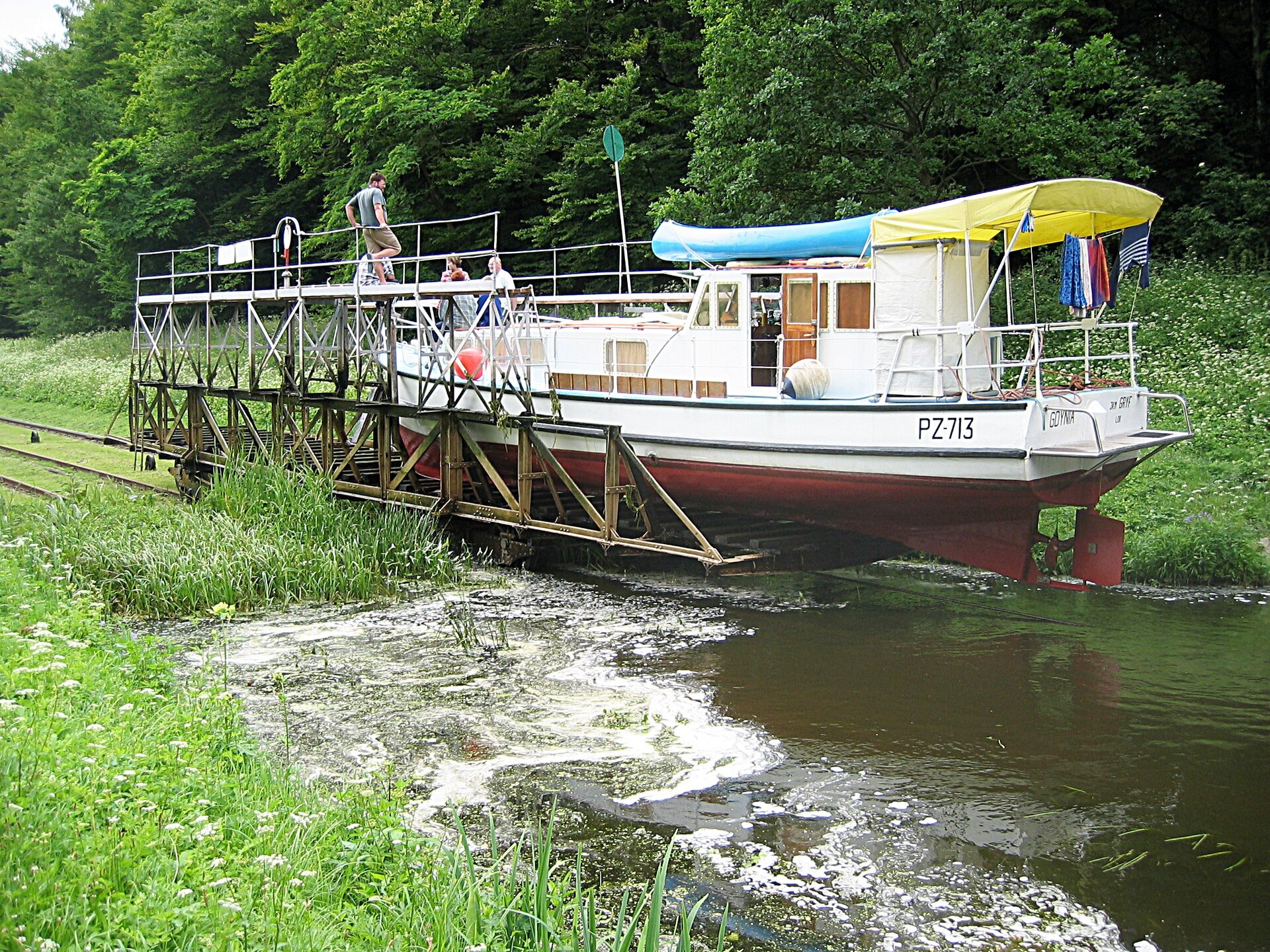 Fotografia przedstawia pochylnię na Kanale Elbląskim. Widać statek umieszczony na platformie, która jedzie po szynach. Szyny znajdują się na lądzie, a platforma w połowie znajduje się na lądzie, a w połowie nad wodą.