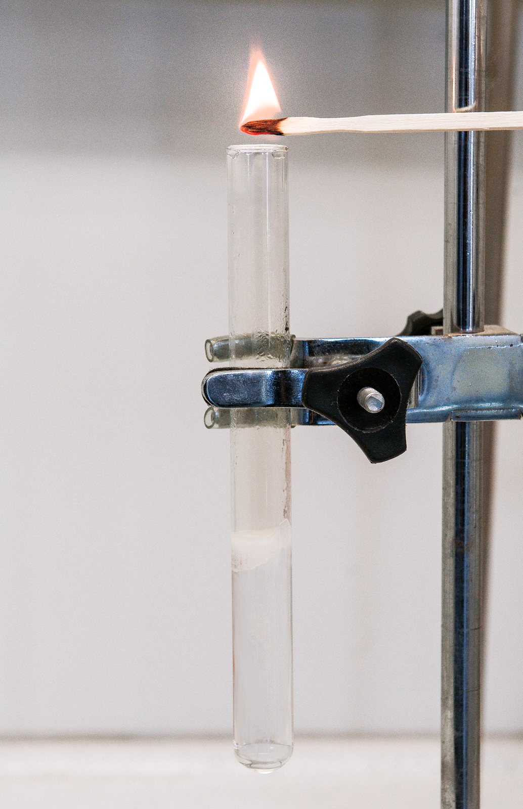 Grafika przedstawia zdjęcie z wykonywania doświadczenia. Na zdjęciu znajduje się szklana przezroczysta probówka zawieszona na metalowym statywie. Nad probówką jest zapalone drewniane łuczywko. Płomień znajduje się nad wylotem z probówki. W jednej trzeciej objętości probówki znajduje się bezbarwna ciecz, która zaczyna się pienić.