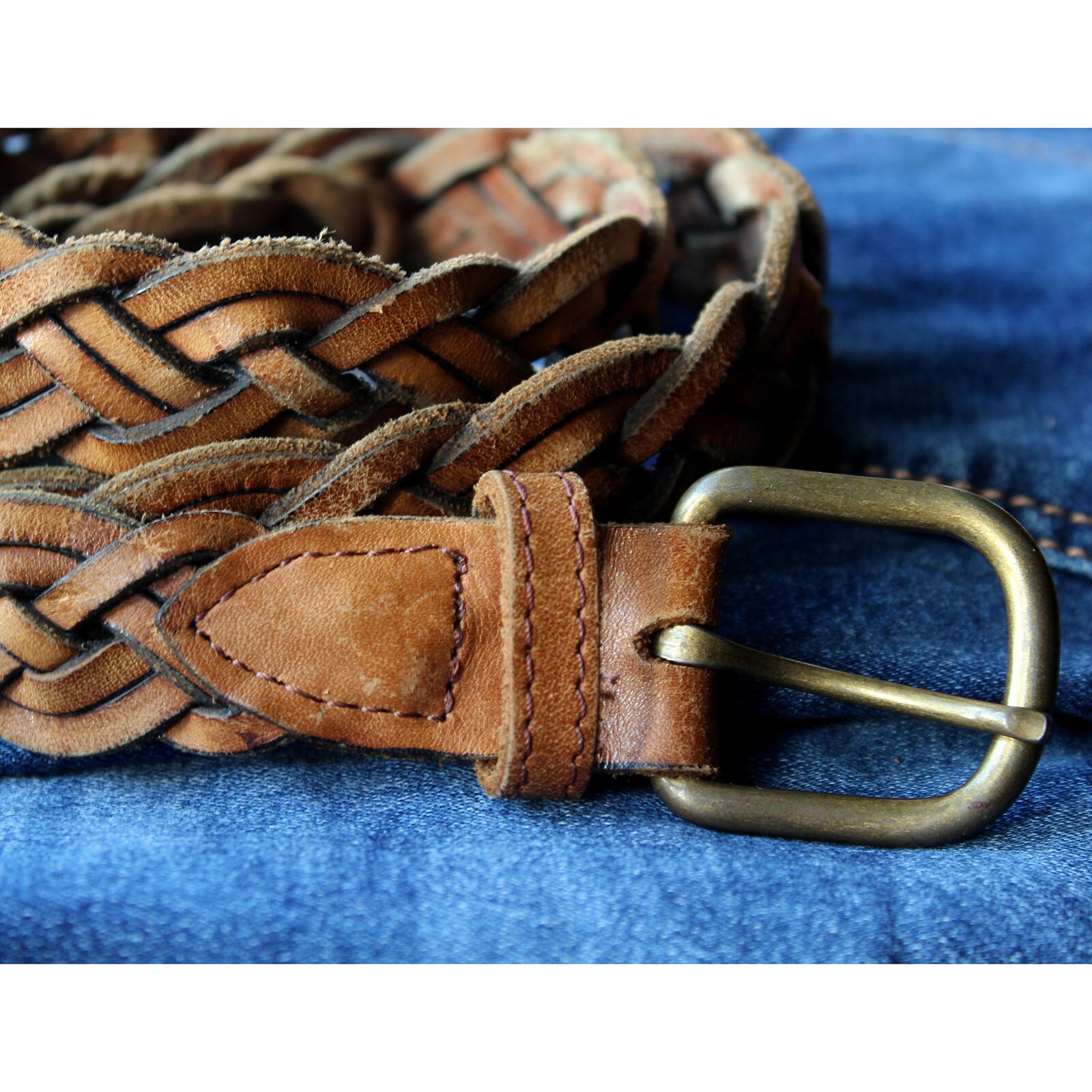Zdjęcie przedstawia brązowy, skórzany pasek leżący na dżinsie. Pasek został wykonany z plecionki w kształcie warkocza i ma kwadratową klamrę z mosiądzu. 
