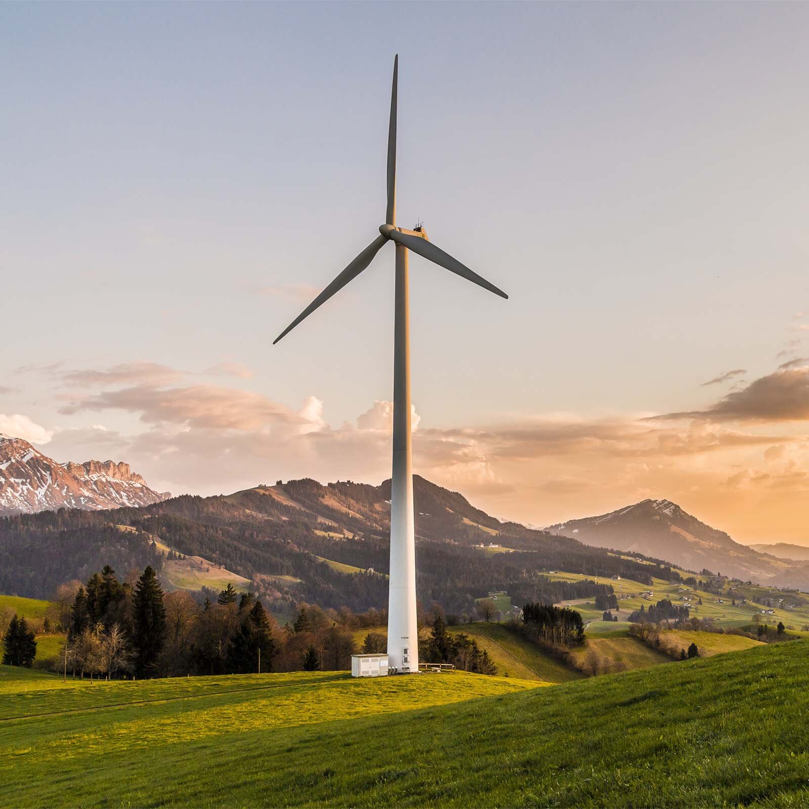 Zdjęcie przedstawia stojący wiatrak w dolinie otoczonej pagórkami. 