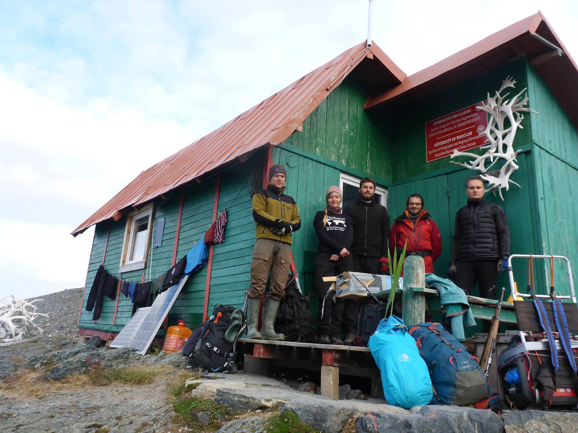 Na zdjęciu grupa badaczy przed budynkiem stacji badawczej. Ciepło ubrani, przed nimi leżą plecaki. Stacja przypomina mały, drewniany domek. 