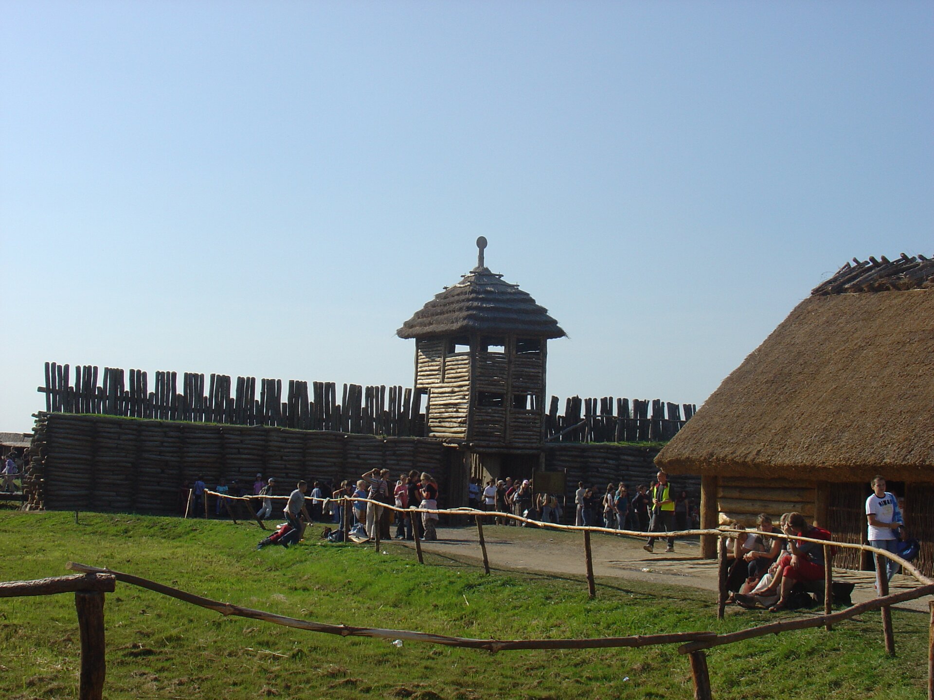 Fotografia przedstawia fragment drewnianej osady. W prawym rogu widać fragment drewnianej chałupy krytej strzechą, za nią wał z bali drewnianych i wieża z otworami. Pod wieżą, zamykane na wrota wejście do osady. Wał chronił mieszkańców przed najazdem wrogów. 