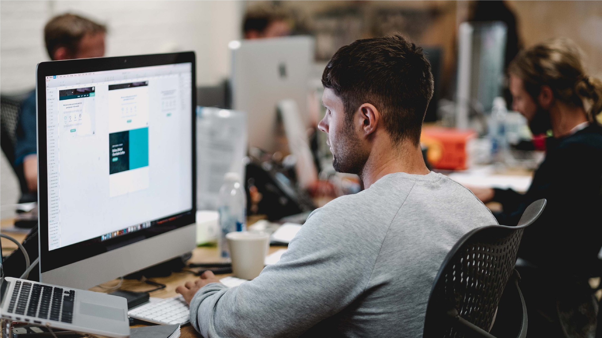 Zdjęcie przedstawia młodego mężczyznę pracującego w biurze. Mężczyzna siedzi przed dużym ekranem komputera. W tle zdjęcie jest trochę rozmyte i widać innych ludzi pracujących w tym biurze.