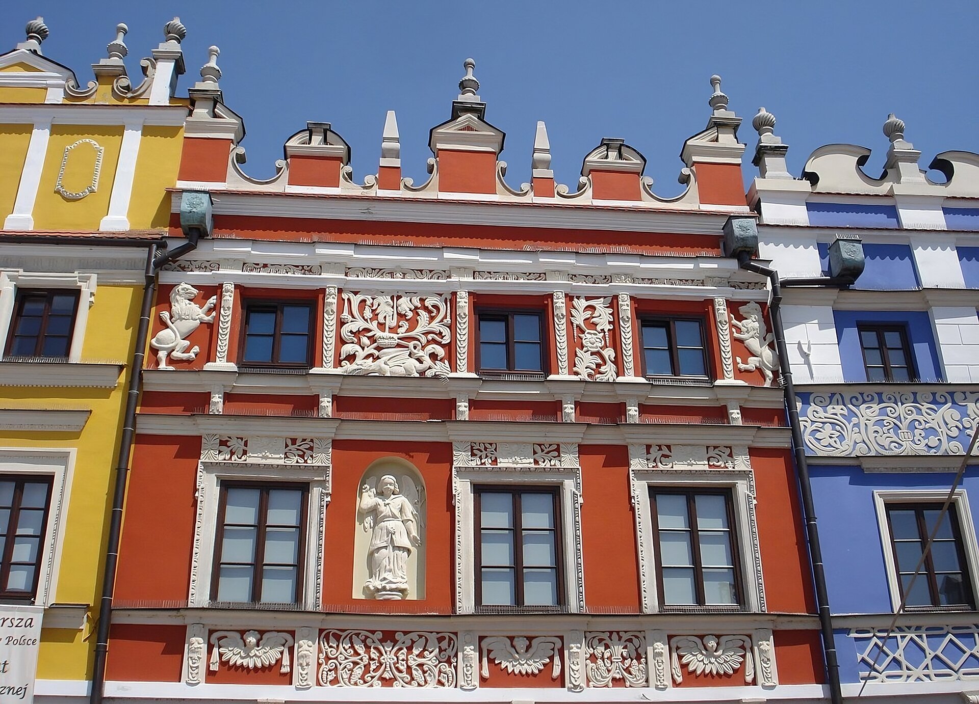 Zdjęcie przedstawia widok współczesny na górną partię jednej z kamienic ormiańskich na rynku w Zamościu. Kamienica o czerwonym kolorze ściany, prostokątnych oknach, jest dzielona kondygnacjami na fasadzie przez gzymsy i fryzy. Na szczycie fasady renesansowa attyka osłaniająca dach, zdobiona sterczynami i wolutami. Dominują zdobnicze motywy roślinne i zwierzęce na fryzach. Pośrodku jednej z kondygnacji, piętra uchwyconego bliżej dolnej krawędzi zdjęcia, widoczna we wnęce figura postaci. Po obu stronach czerwonej kamienicy fragmenty attyk i ścian kamienic sąsiednich - żółtej z lewej strony i błękitnej z prawej.