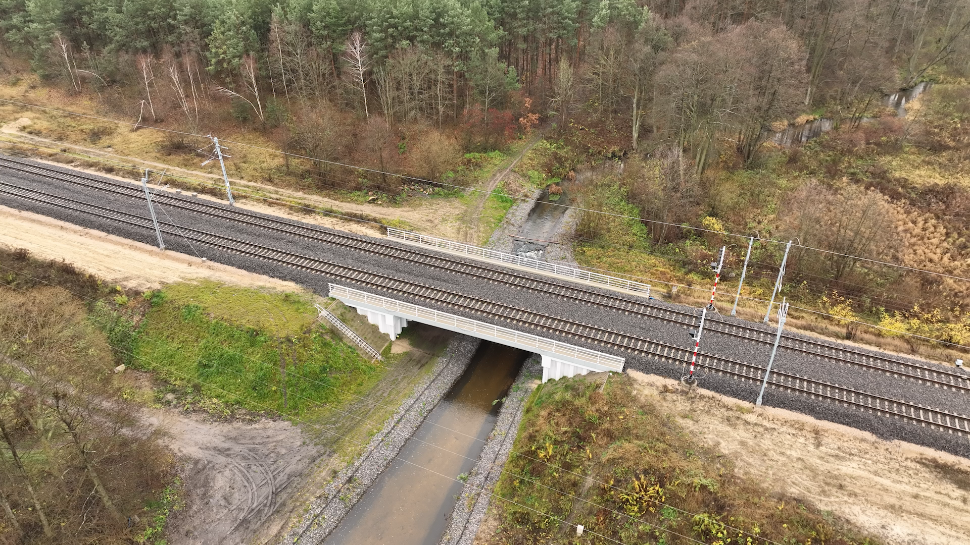  Na zdjęciu widać most belkowy z perspektywy lotu ptaka. Składa się on z dwóch potężnych, betonowych belek i został wybudowany nad rzeką. Tory kolejowe przechodzą przez las. Most belkowy służy do przeprawy pociągu przez rzekę.