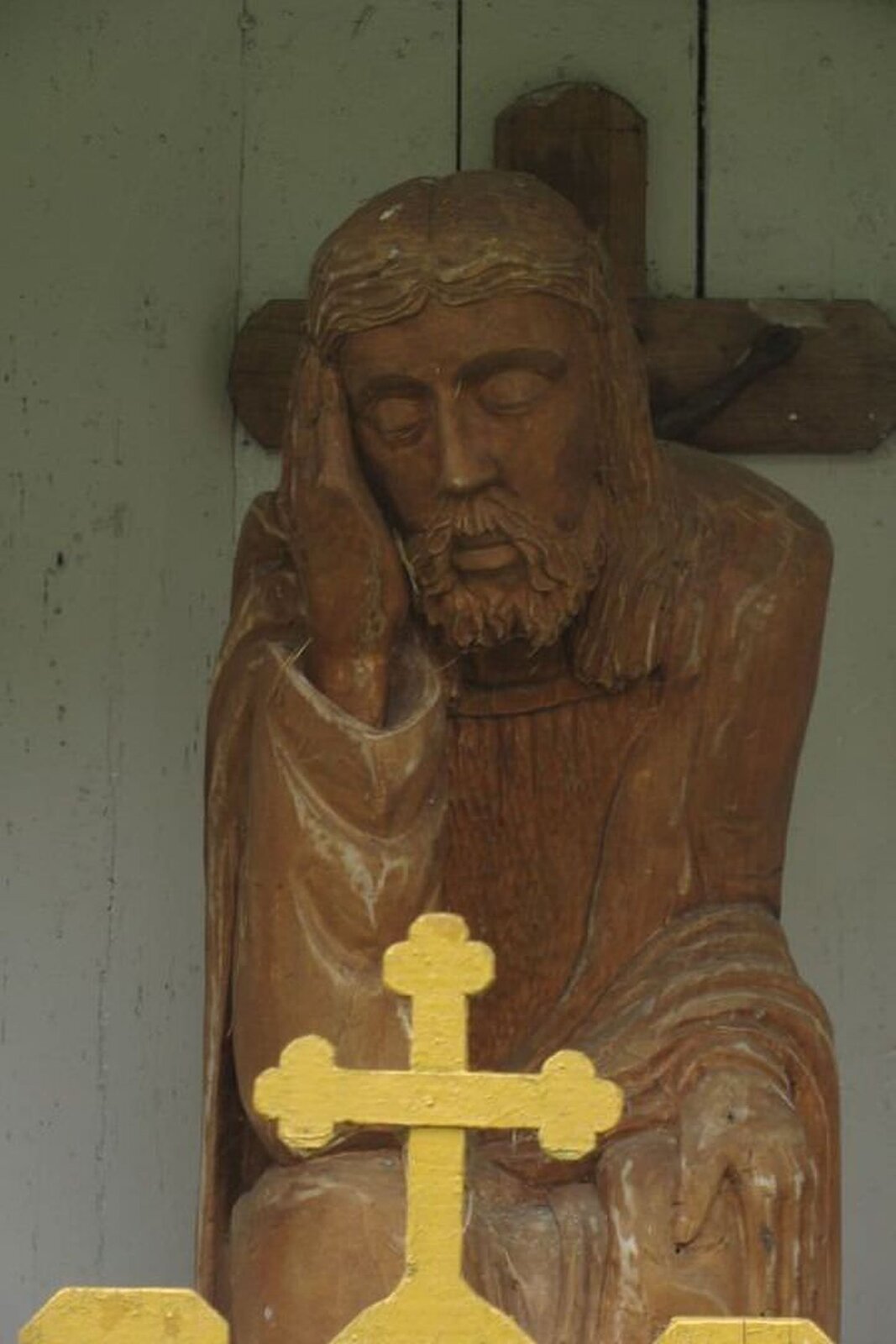 Ilustracja przedstawia drewnianą rzeźbę "Chrystusa frasobliwego". Ukazuje Jezusa z zamkniętymi oczami. Jest ukazany w pozycji siedzącej, prawą ręką podpiera głowę, lekko pochyloną w lewą stronę. Za nim znajduje się krucyfiks zawieszony na białej ścianie z desek.