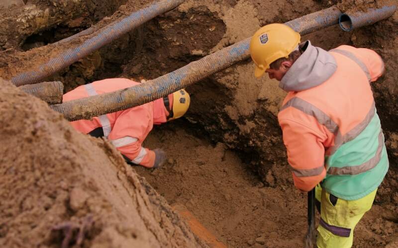 Krok szósty, Prowadzenie renowacji przewodów sieciowych. Fotografia przedstawia dwóch pracowników w wykopie, pracujących przy renowacji przewodów sieciowych. Nad wykopem przewody z grubych rur z tworzywa sztucznego.