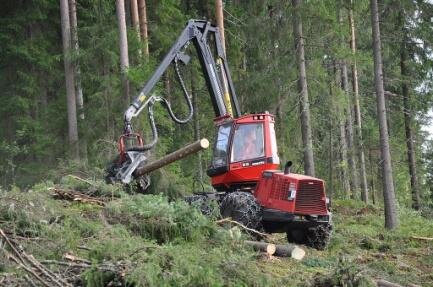 Zdjęcie przedstawia kombajn zrębowy wykorzystywany przy wycince lasu. Na zdjęciu maszyna z wysięgnikiem, która przenosi z miejsca na miejsce ucięte pnie drzew. Wokół leżą ścięte gałęzie drzew. 
