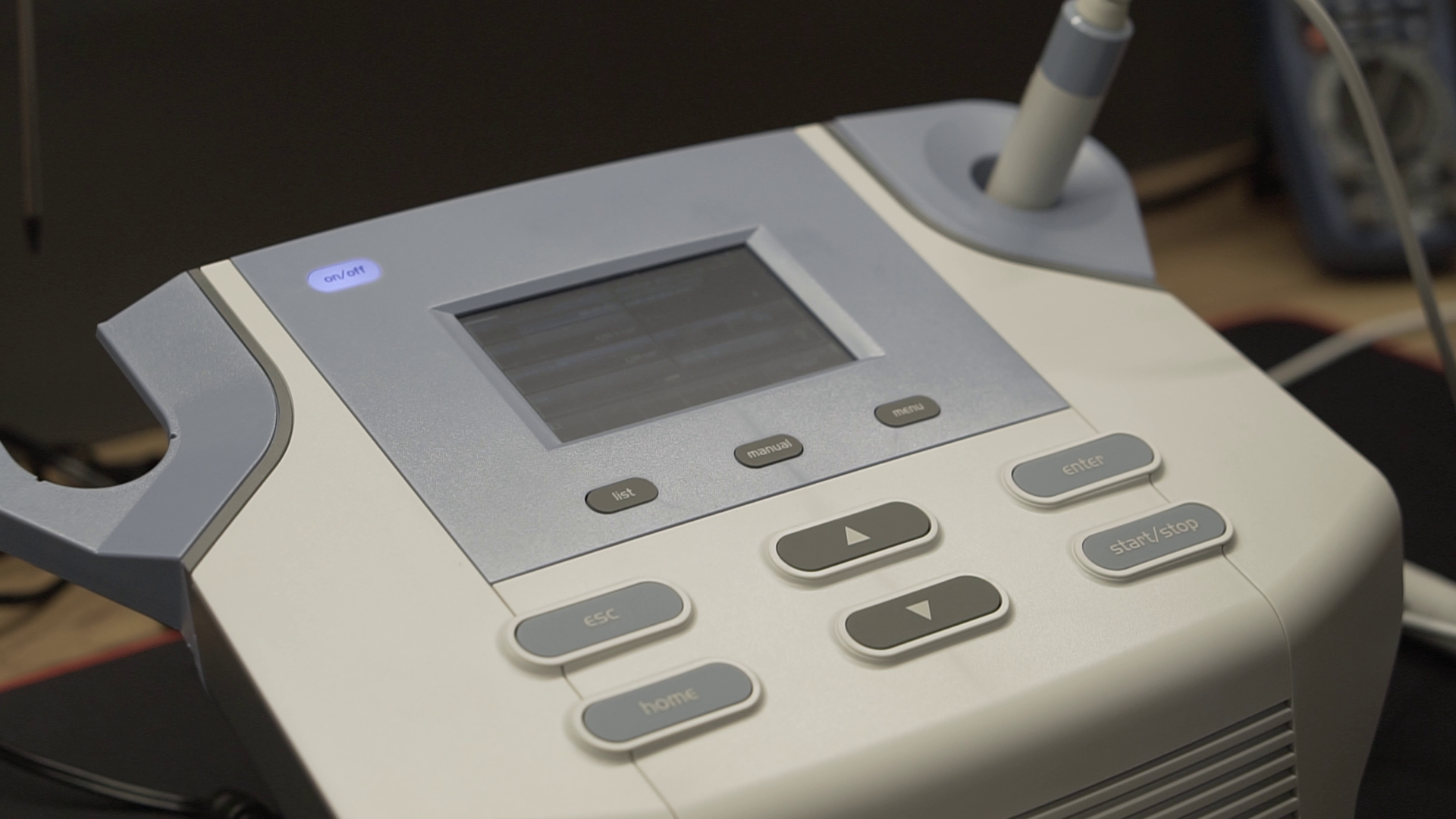 Zdjęcie przedstawia aparat do laseroterapii. Na obudowie korpusu urządzenia znajduje się ekran i przyciski sterujące. Po prawej stronie jest głowica laserowa na przewodzie.