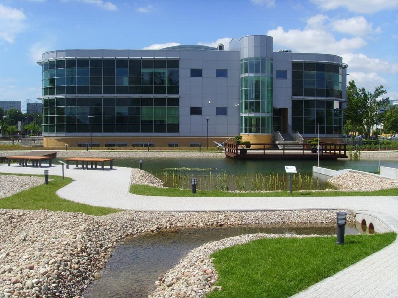 Zdjęcie. W oddali budynek biurowy. Przed budynkiem zbiornik wodny z pomostem. Do zbiornika doprowadzone jest płytkie koryto wyłożone kamieniami, w którym znajduje się woda. Wzdłuż brzegu zbiornika poprowadzony jest chodnik. Na chodniku widoczne są dwie ławki.