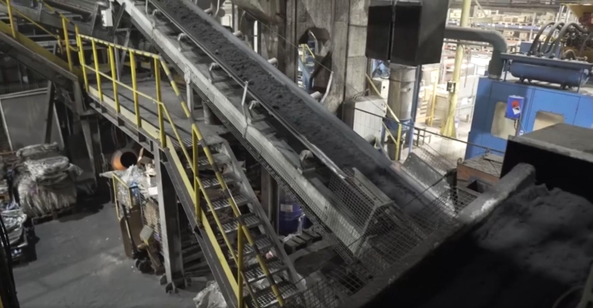 Na zdjęciu widoczna jest linia taśmowa transportująca masę formierską. W centralnej części zdjęcia znajduje się przenośnik. Przenośnik jest zbudowany ze stalowych modułów na których osadzone są rolki. Na rolkach rozciągnięta jest taśma. Obok taśmociągu znajdują się schody. Schody zabezpieczone są barierką. Całość urządzenia znajduje się w dużym budynku przemysłowym. 